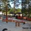 Wippe Robinie auf Spielplatz mit Sandboden und Kiefern im Hintergrund.
