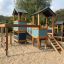 Spielturm mit Klettergerüst, Rutsche und Schaukel auf einem Sandboden im Freien.