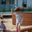 Kindermädchen springt von einem Holzbalken im Spielbereich, während ein kleiner Junge sitzt und zusc