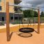 Nestschaukel Robinie auf modernem Spielplatz, im Hintergrund Gebäude und Grünflächen.