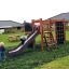 Klettergerüst mit Kletterwand und Rutsche auf einem grünen Rasen mit spielenden Kindern.