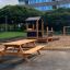 Holz-Spieltisch und Sitzbänke auf einem Spielplatz, umgeben von Grünflächen.