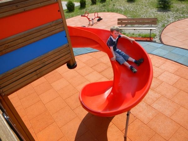 Kind auf roter Rutsche im Spielplatzbereich mit Fallschutzplatten.