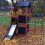 Bastei Spielhaus mit Kletternetz und Rutsche auf einem Herbstblätter bedeckten Spielplatz.