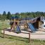 Spielplatz mit Holzspielturm und Rutschen auf Sandboden in natürlicher Umgebung.