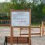 Informationstafel aus Holz mit Spielplatzregeln neben einer Parkbank auf sandigem Boden.