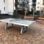 Freistehende Betontischtennisplatte auf einem gepflasterten Platz mit Bänken im Hintergrund.