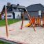 Doppelter Schaukel Rahmen auf einem Spielplatz mit Sandboden und Rutsche im Hintergrund.