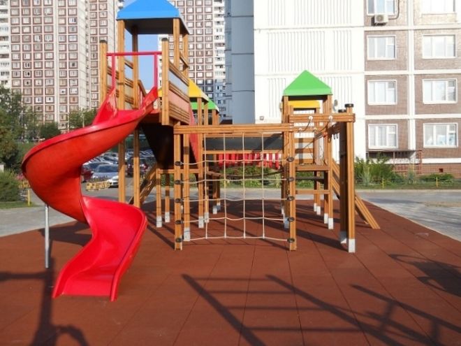 Spielplatzgerät mit Rutsche, Kletterturm und Brücke, im Hintergrund Mehrfamilienhäuser.