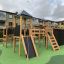 Holzspielgerät mit Tunnel, Kletterwand, Rutsche und Türmchen auf einem Spielplatz.