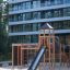 Spieltürme mit Tunnel, Kletterwand und Rutsche aus Holz auf einem Spielplatz vor modernem Gebäude.
