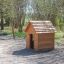 Holzspielhaus Natur Spielhaus im Freien auf einem Spielplatz mit Schaukel im Hintergrund.